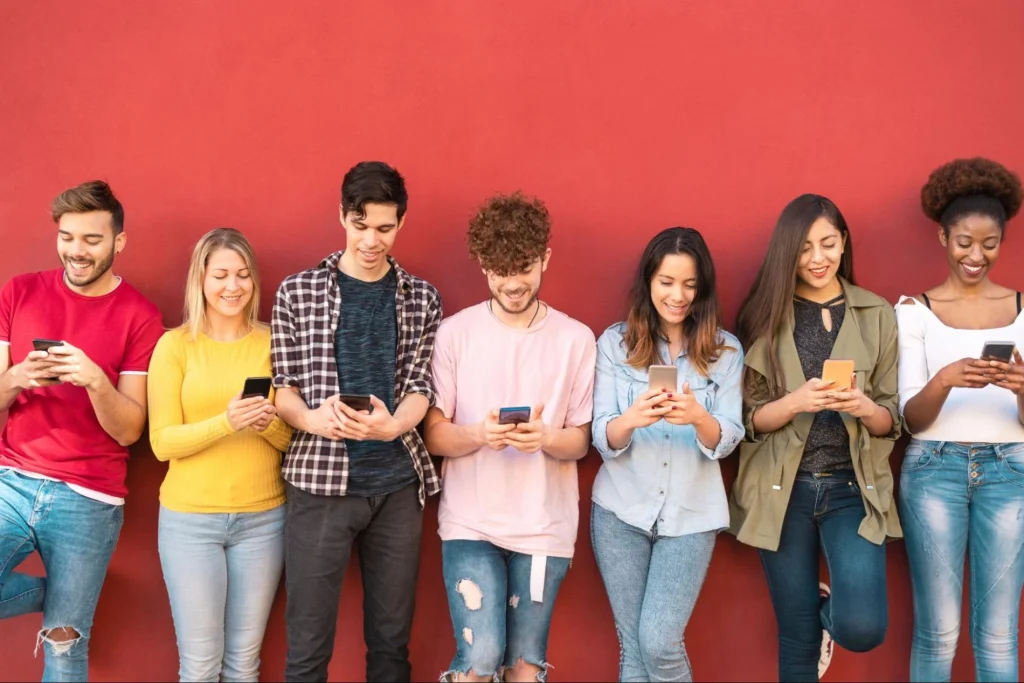 Group of young adults using smartphones while standing against a red wall, representing social media engagement and digital communication trends.